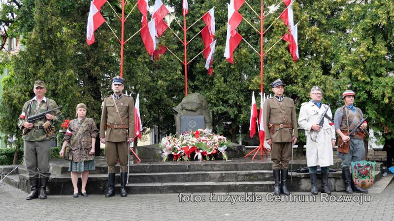 W centrum znajduje się pomnik na tle drzew, a pod nim kwiaty. Nad  pomnikiem stoją trzy słupy z biało-czerwonymi flagami. Po lewej i prawej stronie stoją ludzie ubrani w wojskowe mundury.