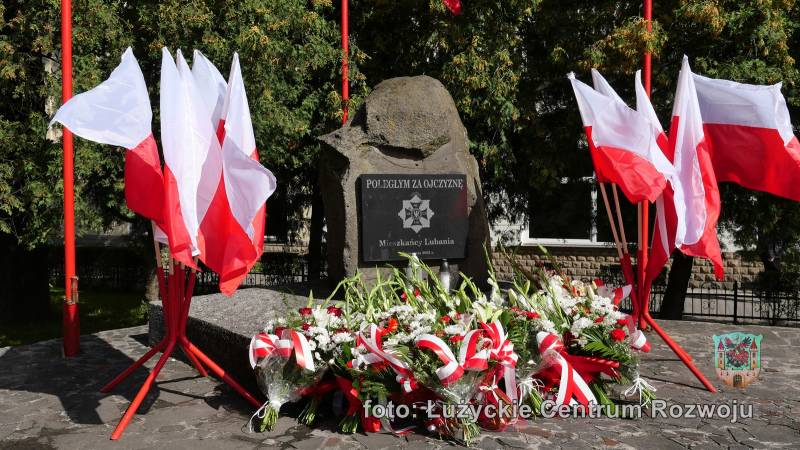 W centrum znajduje się pomnik. Pod nim stoją kwiaty. Po obu stronach pomniku stoją flagi Polski.