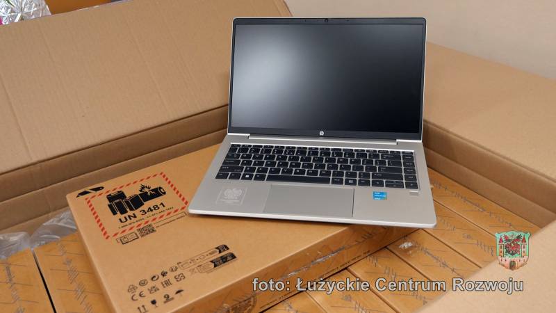 Laptop z srebrną obudową i czarnymi klawiszami, stoi na jasnobrązowym pudełku.