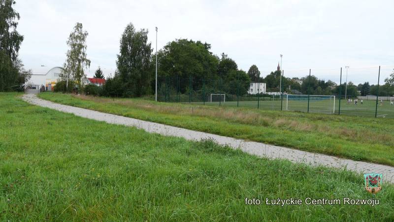 Trawnik przedzielony przez chodnik z kostki prowadzący do kładki. W tle drzewa i ogrodzone siatką boisko do piłki nożnej.