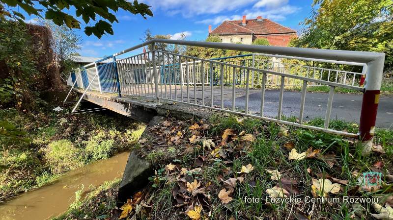 Widok na barierkę od mostu, na pierwsym planie trawa z jesiennymi liśćmi. Pod mostem płynie rzeka, w tle budynek.